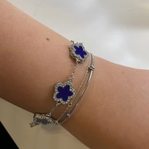 Silverarmband med blå blommor - Elegant silverarmband med flera blå blomformade hängen. Varje blomma är omgiven av en glittrande kant som ger en lyxig känsla. Perfekt för att lägga till en färgklick till din outfit.
