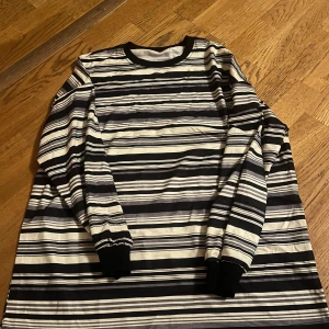 Randig långärmad tröja - Säljer en stilren randig tröja med långa ärmar. Tröjan har ett klassiskt mönster i svart, vit och beige med ribbade muddar vid ärmslut och hals. Perfekt för en avslappnad look.