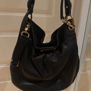Svart axelväska från Marc by Marc Jacobs - Snygg svart axelväska i skinn från Marc by marc bacobs med guldfärgade metalldetaljer. Passar bra som skolväska! Väskan har en justerbar axelrem och en stilren design med en metallplatta framtill. Den är köpt 2000kr och säljer därför för halva priset (1000kr). Vintage. Skriv vid intresse, pris kan diskuteras🥰