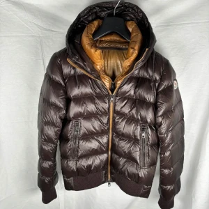 Moncler jacka  - (Jackan är köpt på Store Enda Ab) Säljer för endast 3499kr sommarpris.Snygg brun dunjacka från Moncler med huva och dragkedja. Jackan har en quiltad design och ribbade muddar vid ärmslut och nederkant. Perfekt för kyliga dagar. Västen inuti går även att ta av. Passar någon som är 175cm och under.
