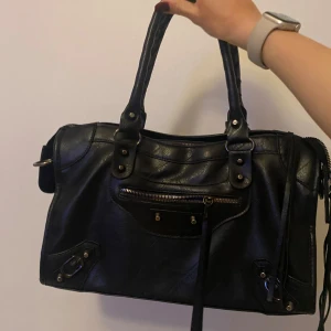 Handväska - Snygg svart handväska i skinn med coola detaljer. Väskan har en praktisk dragkedja och nitar som ger en edgy look. Perfekt för att bära dina dagliga nödvändigheter med stil.
