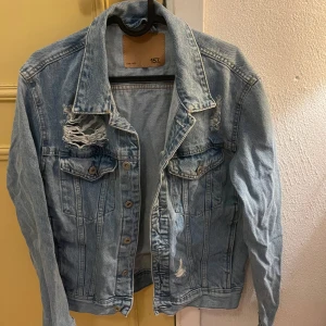 Ljusblå jeansjacka - Snygg ljusblå jeansjacka med slitna detaljer och knappar framtill. Jackan har två bröstfickor med lock och en klassisk krage. Perfekt för en avslappnad stil.