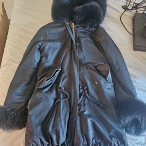 Svart parkajacka med pälsdetaljer - Snygg svart parkajacka med fluffig päls på huva och ärmslut. Jackan har dragkedja och knappar framtill samt praktiska fickor med knappning. 