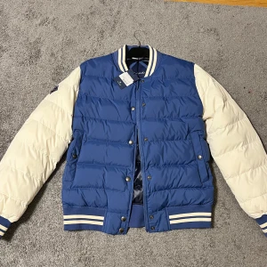Blå och vit pufferjacka från GANT - Snygg blå och vit pufferjacka från GANT med ribbade muddar och krage. Jackan har knappar framtill och ett broderat märke på ärmen. Ord. pris 2499. Perfekt för kyligare dagar. 
