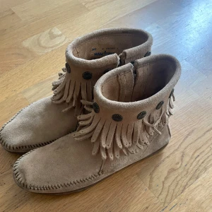 Beige mocka boots från Minnetonka - Säljer ett par beige mocka boots från Minnetonka med fransar och dekorativa knappar. Perfekta för en bohemisk stil. De har en dragkedja på sidan för enkel på- och avtagning.