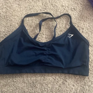 Mörkblå sporttopp från Gymshark - Säljer en mörkblå sporttopp från Gymshark med smala axelband och en stilren design. Perfekt för träning och ger bra stöd. Toppen har en liten logga på framsidan och korsade band i ryggen.