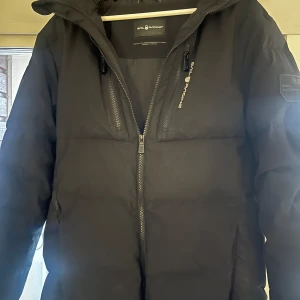 Sail Racing Jacks - Snygg svart dunjacka från Sail Racing med dragkedja och hög krage. Jackan har långa ärmar och två bröstfickor.