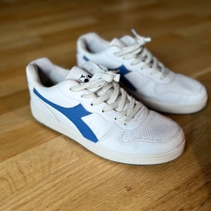 Vita och blå sneakers från Diadora - Snygga vita sneakers från Diadora med blå detaljer och snörning. Perfekta för en avslappnad stil. Skorna har en klassisk design med perforerade detaljer på ovandelen.