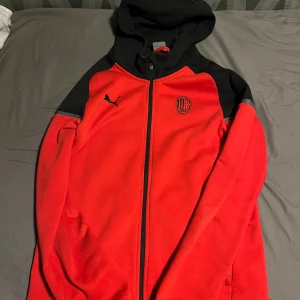 Röd och svart huvtröja från Puma/Milan - Snygg röd och svart huvtröja från Puma med AC Milan-logga på bröstet. Tröjan har dragkedja och huva, perfekt för fotbollsfans. Den är långärmad och har en sportig look. Nyskick.