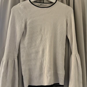 Vit ribbad tröja från Gina Tricot - Säljer en stilren vit ribbad tröja från Gina Tricot. Tröjan har långa ärmar med volangdetaljer och en svart kant vid halsringningen. Perfekt för en chic och bekväm look.