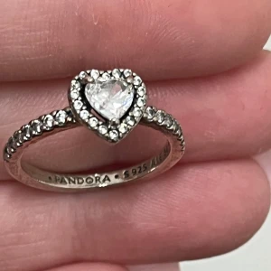 Silverring med hjärtformad sten från Pandora - Elegant silverring från Pandora med en gnistrande hjärtformad sten i mitten, omgiven av små glittrande stenar. Perfekt för att ge en touch av glamour till vilken outfit som helst.