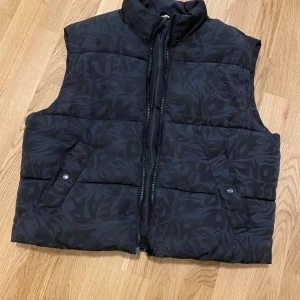 Svart dunväst med mönster No fear x H&M jacket size m  - Snygg svart dunväst från Zara med diskret mönster. Västen har dragkedja och två fickor med knappar framtill. Perfekt för kyligare dagar när du vill hålla stilen.