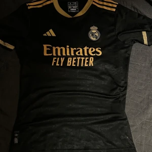 Svart Real Madrid fotbollströja - Snygg svart Real Madrid tröja från Adidas med guldiga detaljer och klubbens emblem på bröstet. Kortärmad design med sponsorlogga 'Emirates Fly Better' i guld. Perfekt för fans som vill visa sitt stöd med stil.                        B: 45 L: 65