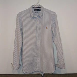 Randig skjorta från Ralph Lauren - Snygg långärmad skjorta från Ralph Lauren i blått och vitt randigt mönster. Skjortan har en klassisk krage och knappar framtill. Den är broderad med det ikoniska Ralph Lauren-logotypen på bröstet. Skjortan är i nyskick och varsamt använt. Prisen kan vara diskuterbart vid snabb affär. Bara att höra av sig kring funderingar.