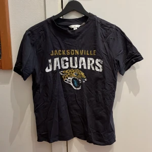 Svart Jacksonville Jaguars t-shirt från H&M - Snygg svart t-shirt från H&M med Jacksonville Jaguars tryck i gult och vitt. T-shirten är kortärmad och har en klassisk rund halsringning. Säljer för att jag har bytt stil.