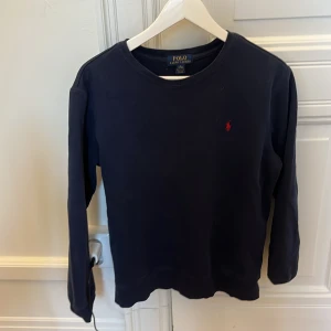 Mörkblå tröja från Polo Ralph Lauren - Säljer en stilren mörkblå tröja från Polo Ralph Lauren med rund halsringning och ett diskret rött logotypbroderi på bröstet. Tröjan har långa ärmar och är perfekt för en avslappnad men elegant look.
