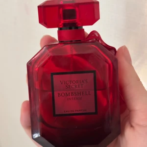 Bombshell Intense Eau de Parfum från Victoria's Secret - Säljer en elegant röd parfymflaska från Victoria's Secret, Bombshell Intense. Flaskan har en facetterad design med en matchande röd kork och ett dekorativt band runt halsen. Perfekt för den som älskar en intensiv och förförisk doft.