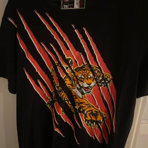 Svart t-shirt med tigertryck från Plein Sport - Cool svart t-shirt från Plein Sport med ett stort tigertryck i orange och rött. Perfekt för en djärv stil. T-shirten har korta ärmar och en normal passform.