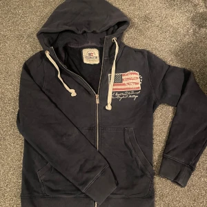 Mörkblå ziphoodie från Lexington - Säljer en riktigt fet mörkblå hoodie från Lexington med dragkedja och tryck av en amerikansk flagga på bröstet. Tröjan har en justerbar huva med snören och långa ärmar. 
