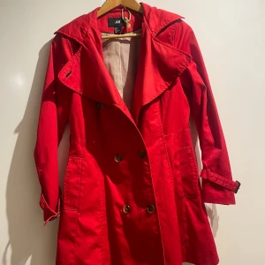 Röd trenchcoat från H&M - Snygg röd trenchcoat från H&M med dubbelknäppning och bred krage. Jackan har långa ärmar och ett bälte i midjan för en justerbar passform. Perfekt för höst och vår! Har tyvärr tappat bort bältet. Priset går att diskutera! 