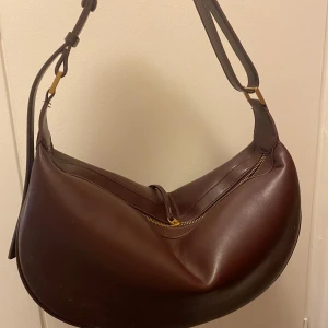 Väska från Zara - Snygg mörkröd/vinröd axelväska med justerbar axelrem och dragkedja. Perfekt för att bära dina dagliga nödvändigheter med stil.