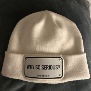 Vit mössa från John Hatter & Co - Vit mössa med uppvikt kant och en metallplatta framtill med texten 'WHY SO SERIOUS?'. Perfekt för att hålla stilen och värmen under kyliga dagar.