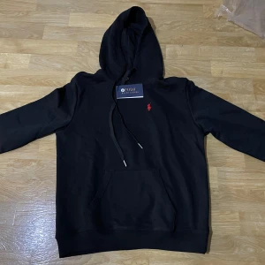 Svart hoodie från Polo Ralph Lauren - Säljer en stilren svart hoodie från Polo Ralph Lauren med en liten röd logga på bröstet. Den har en klassisk känguruficka och justerbar huva med snören. Perfekt för en avslappnad look.