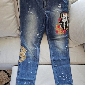 Blå jeans med broderade detaljer - Unika blå jeans med måladeblommor i gult, brunt och rött samt en tryckt design av en figur med kors. Jeansen har en sliten look med vita färgstänk och är dekorerade på både fram- och baksida. Perfekt för dig som vill sticka ut!
