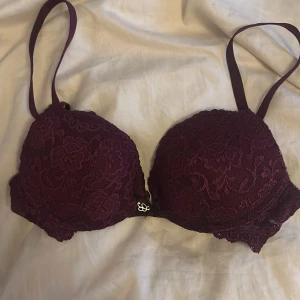 Vinröd spets- push up bh - Säljer en elegant vinröd bh med spetsdetaljer från Cubus. Den har justerbara axelband och en liten dekorativ detalj framtill. Perfekt för att känna sig extra fin under kläderna.