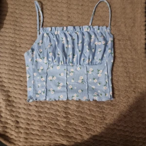 Blommig blå linnetopp - Söt blå linnetopp med små blommor i vitt och rosa. Toppen har smala axelband, croppad och en ribbad struktur. Perfekt för varma sommardagar!