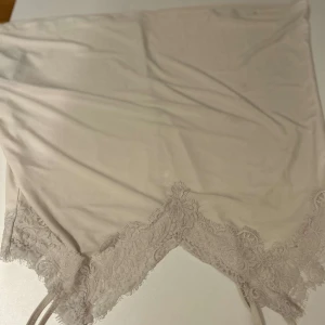 Vit axelbandslös topp med spetsdetaljer - Säljer en elegant vit axelbandslös topp med vacker spetsdetalj längs nederkanten. Perfekt för en stilren och chic look. Toppen är tillverkad i ett mjukt och följsamt material.