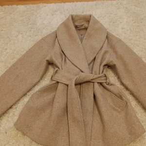 Beige kappa från Gina Tricot - Säljer en stilren beige kappa från Gina Tricot i premiumkvalitet. Den har en elegant knytning i midjan och långa ärmar. Perfekt för höst och vår med sin klassiska design och mjuka material. Använd 2-3 gånger. 
