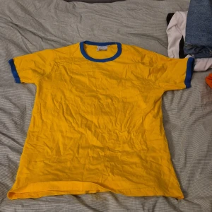 Gul t-shirt med blå detaljer från The Big Apple - Säljer en gul t-shirt med blå krage och ärmslut från The Big Apple. Perfekt för en avslappnad stil. 🟡🔵