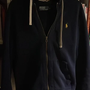 Mörkblå zip-hoodie från Ralph Lauren - Säljer en snygg mörkblå hoodie från Ralph Lauren med dragkedja och vit snörning. Den har en broderad gul logotyp på bröstet. Perfekt för en stilren och avslappnad look. Använd fåtal gånger. Priset inte hugget i sten