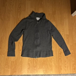 Grå full zip från These Glory Days - Säljer en stilren full zip hoodie från These Glory Days i mjukt material. Tröjan har en dragkedja framtill och långa ärmar, perfekt för kyligare dagar. Den är enkel att matcha med olika outfits och ger en avslappnad look. Modellen är 169cm och väger 52kg. Tröjan är köpt från volt för 1100kr.