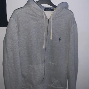 Zip tröja Ralph Lauren - Säljer en klassisk grå hoodie från Ralph Lauren med dragkedja och huva. Den har en liten broderad logga på bröstet och snören vid huvan. Perfekt för en avslappnad stil.