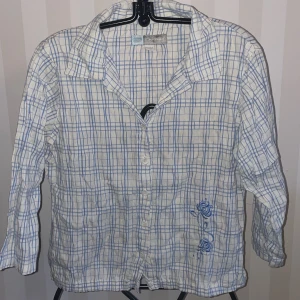 Vit och blå rutig skjorta från Drop st.158-164 - Säljer en vit och blå rutig skjorta från Drop med broderade blommor i blått på framsidan st158-164. Skjortan har långa ärmar och knappar framtill. Perfekt för en avslappnad stil med en touch av detaljer.