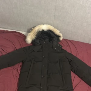 Svart dunjacka från Canada Goose  - Säljer en svart dunjacka från Canada Goose med avtagbar päls på huvan. Jackan har långa ärmar och knäppning med både dragkedja och knappar. Perfekt för kalla vinterdagar.