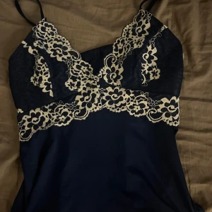 Mörkblått linne med spetsdetaljer - Elegant mörkblått linne med vackra spetsdetaljer i vit. Linnet har smala axelband och en mjuk, följsam passform. Perfekt för en stilfull look.