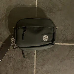 Svart axelväska från Stone Island - Snygg svart axelväska från Stone Island med dragkedja och logotyp på framsidan. Perfekt för att bära med sig det viktigaste. Stilren design som passar till många tillfällen.