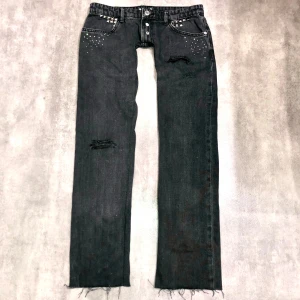Svarta 2000s jeans med nitar och slitningar - Coola svarta jeans med nitar vid fickorna och slitningar på benen. Jeansen är lågmidjade och har bootcut passform. De är dekorerade med små glittriga rhineatones detaljer både fram och bak. Perfekta för en edgy look! OBS de är extremt, EXTREMT lågmidjade! Skriv för mått.