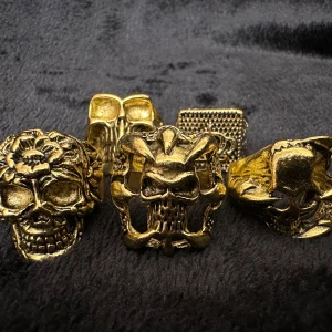 Fem feta guldiga ringar med dödskallemotiv  - Säljer ett set med fem feta guldiga ringar med dödskallemotiv. Varje ring har en distinkt design med detaljer som blommor och korslagda ben. Olika storlekar för att passa olika fingrar. Mellan 1,8 och 2,5 cm i innerdiameter. Perfekt för att ge en edgy touch till din stil. Nya och oanvända. 