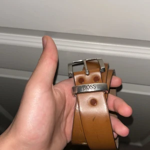 Brunt skärp från Boss - Snyggt brunt skärp från Boss i skinn med en stilren silverfärgad spänne. Perfekt för att ge en klassisk touch till din outfit. Skärpet har flera hål för justerbar passform.