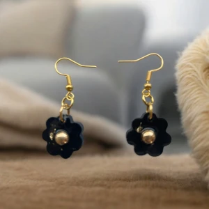 Blommiga örhängen med gulddetaljer - Charmiga örhängen i form av svarta blommor med en guldfärgad mitt. De har en elegant krok i guldton som ger en fin kontrast till den mörka blomman. Perfekta för att lägga till en touch av stil till din outfit.