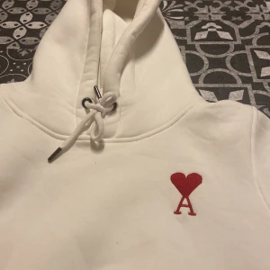 Vit hoodie med rött hjärta från Ami - Säljer en stilren vit hoodie från Ami med ett broderat rött hjärta och bokstaven 'A' på bröstet. Hoodien har en klassisk design med dragsko i huvan och en stor ficka framtill. Perfekt för en avslappnad look.