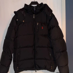 Svart dunjacka från Polo Ralph Lauren - Säljer en svart dunjacka från Polo Ralph Lauren med dragkedja och huva. Jackan har en liten röd logga på bröstet och praktiska fickor med dragkedjor. Perfekt för kyliga dagar.