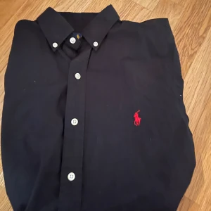 Svart skjorta från Ralph Lauren - Säljer en stilren svart skjorta från Ralph Lauren i slim fit. Skjortan är tillverkad av 100% bomull och har en klassisk krage med knappar. 