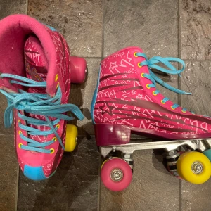 Färgglada rullskridskor med stjärnmönster - Säljer ett par färgglada rullskridskor i rosa med stjärnmönster. De har turkosa snören och gula hjul, vilket ger en lekfull look. Perfekta för en rolig åktur!