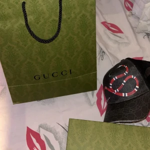 Svart keps från Gucci - Snygg svart keps från Gucci med ett diskret mönster och broderade detaljer i rött och vitt. Kepsen har en justerbar rem baktill för perfekt passform och är tillverkad i ett lätt och luftigt material.
