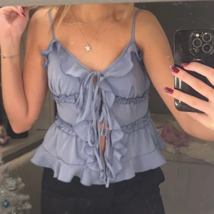Ljusblå volangtopp med knytning - Säljer en söt ljusblå topp med volanger och knytning framtill. Toppen har smala axelband och en luftig passform, perfekt för varma dagar. Den har en romantisk och lekfull stil som passar till många tillfällen, både fest och sommardagar utomhus⭐️ Nypris 350kr, aldrig använd.